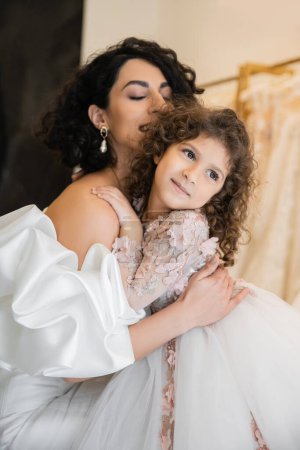 enchanting middle eastern bride with brunette hair and closed eyes in white wedding dress with puff sleeves and ruffles hugging daughter looking away in bridal store, special occasion 