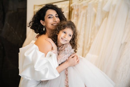 charming middle eastern bride with brunette hair in white wedding dress with puff sleeves and ruffles embracing positive daughter in bridal store, white tulle fabrics on blurred background 