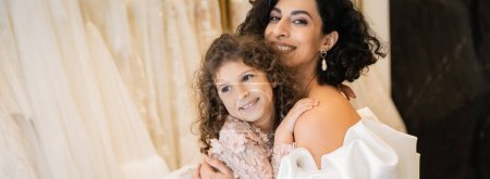 gorgeous middle eastern bride with brunette hair in white wedding dress with puff sleeves and ruffles embracing positive daughter in bridal store, white tulle fabrics on blurred background, banner 