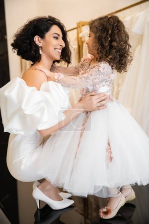 happy middle eastern bride in white wedding dress with puff sleeves and ruffles embracing cheerful little daughter in bridal store, white tulle fabrics on blurred background, high heels