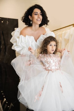 captivating middle eastern bride with brunette hair standing in white wedding gown with puff sleeves and ruffles and looking away while holding tulle skirt of surprised daughter in bridal store 