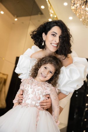 Charmante Braut aus dem Mittleren Osten mit brünetten Haaren steht im weißen Brautkleid mit Puffärmeln und Rüschen hinter der niedlichen Tochter und lächelt zusammen im Brautladen in die Kamera 