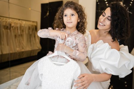 charming middle eastern bride with brunette hair standing in white wedding gown with puff sleeves and ruffles looking at cute daughter holding girly dress with tulle skirt in bridal boutique  