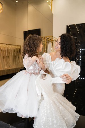 gorgeous middle eastern bride with brunette hair standing in white wedding gown with puff sleeves and ruffles near cute daughter holding girly dress with tulle skirt in bridal boutique  