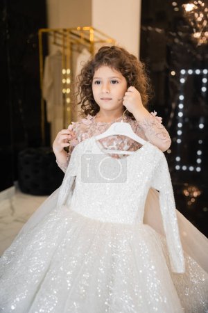 curly middle eastern flower girl in attire holding hanger with white girly dress with tulle skirt in bridal boutique, preparation for wedding, blurred background, golden rack, choosing dress