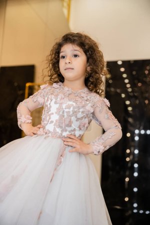 Photo for Curly middle eastern and little girl in floral dress with tulle skirt standing with hands on hips and looking away in bridal boutique, preparation for wedding, blurred background, golden accents - Royalty Free Image