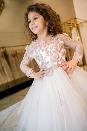 Photo for Joyous middle eastern kid in floral dress with tulle skirt standing with hands on hips and looking away in bridal boutique, preparation for wedding, blurred background, low angle, flower girl - Royalty Free Image