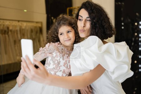 mittelöstliche Braut mit brünetten Haaren in weißem Brautkleid mit Puffärmeln und Rüschen macht Selfie auf Smartphone, während schmollende Lippen neben glücklicher Tochter im Brautladen 