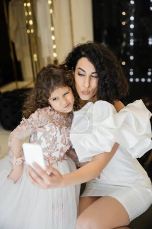 Lustige Braut aus dem Mittleren Osten mit brünetten Haaren in weißem Brautkleid mit Puffärmeln und Rüschen, die ein Selfie auf dem Smartphone mit ihrer Tochter macht, während sie im Brautladen schmollende Lippen macht 