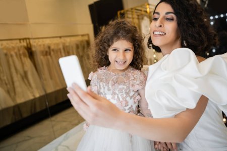charming middle eastern bride with brunette hair in white wedding dress with puff sleeves and ruffles taking selfie on smartphone with happy daughter in bridal store with blurred background 