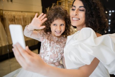 Charmante Braut aus dem Nahen Osten mit brünetten Haaren in weißem Hochzeitskleid mit Puffärmeln und Rüschen, Smartphone in der Hand haltend, während glückliche Tochter beim Videoanruf im Brautladen winkt 