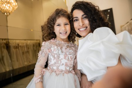 fröhliches kleines Mädchen in floraler Kleidung lächelt neben charmanter Mutter im weißen Hochzeitskleid mit Puffärmeln und Rüschen, während sie gemeinsam in die Kamera in der Brautboutique guckt, Selfie 