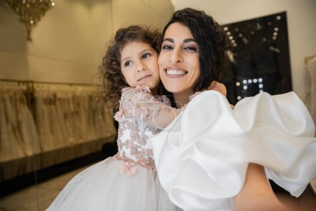 fröhliches kleines Mädchen in floraler Kleidung umarmt ihre charmante Mutter im weißen Hochzeitskleid mit Puffärmeln und Rüschen, während sie in der Brautboutique lächelt und in die Kamera blickt 