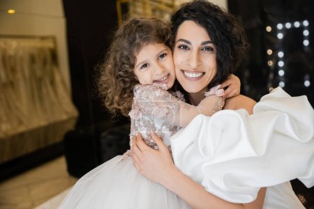 glückliches kleines Mädchen in floraler Kleidung umarmt ihre charmante Mutter im weißen Hochzeitskleid mit Puffärmeln und Rüschen, während sie lächelt und gemeinsam in die Kamera in der Brautboutique schaut 