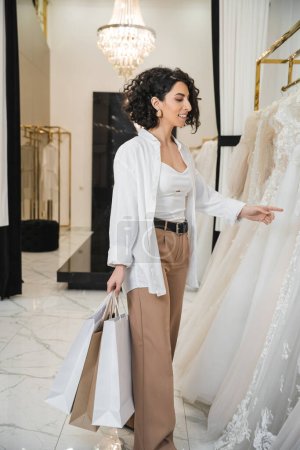 stylische und glückliche Frau aus dem Mittleren Osten mit brünetten und welligen Haaren, die in einer beigen Hose mit weißem Hemd steht und Einkaufstaschen hält, während sie im Brautsalon das Brautkleid auswählt 