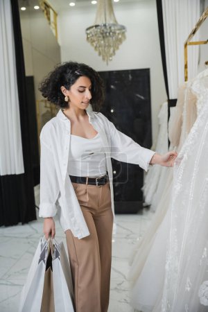 encantadora mujer de Oriente Medio con cabello moreno y ondulado de pie en pantalones de color beige con camisa blanca y la celebración de bolsas de compras al elegir vestido de novia en el salón de bodas, juerga de compras 