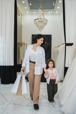 glückliche Frau aus dem Nahen Osten mit brünetten Haaren in beiger Hose und weißem Hemd mit Einkaufstaschen beim Spaziergang mit kleiner Tochter in der Nähe von Brautkleidern im Brautsalon, moderne Braut, Mutter