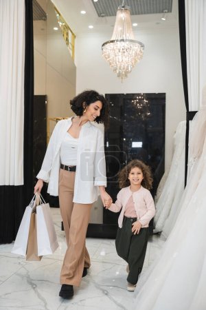 heureuse femme du Moyen-Orient avec des cheveux bruns en pantalon beige avec chemise blanche tenant des sacs à provisions tout en marchant avec fille joyeuse près des robes de mariée dans le salon de mariée, mariée moderne, mère