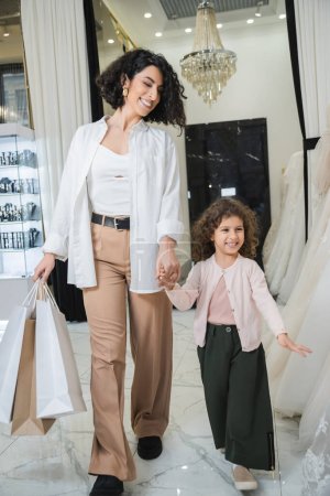 fröhliche Frau aus dem Nahen Osten mit brünetten Haaren in beiger Hose und weißem Hemd mit Einkaufstaschen beim Spaziergang mit kleiner Tochter in der Nähe von Brautkleidern im Brautsalon, moderne Braut, Mutter