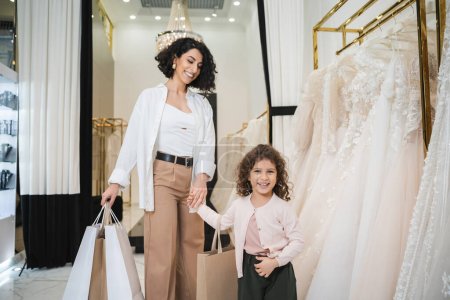 Photo for Happy middle eastern bride with brunette hair in beige pants with white shirt holding shopping bags while standing with little girl near wedding dresses in bridal salon, mother and daughter - Royalty Free Image