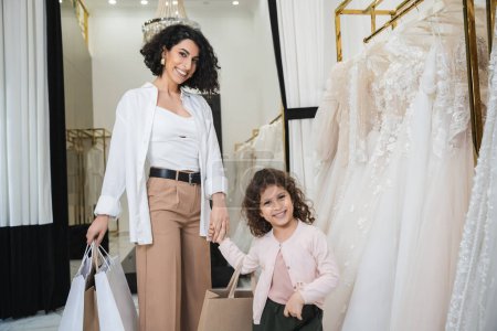 cheerful middle eastern bride with brunette hair in beige pants with white shirt holding shopping bags while standing with little girl near wedding dresses in bridal salon, mother and daughter 