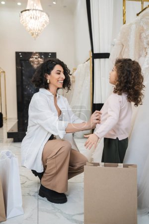 cheerful middle eastern woman with brunette hair in white shirt sitting near shopping bags and wearing cardigan on little girl near wedding dresses in bridal salon, mother and daughter, bride 