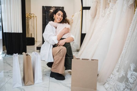 little girl hugging tight cheerful middle eastern bride with brunette hair in white shirt sitting near shopping bags and wedding dresses in bridal salon, mother and daughter, bonding 