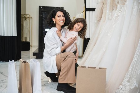Photo for Happy little girl hugging cheerful middle eastern bride with brunette hair in white shirt sitting near shopping bags and wedding dresses in bridal salon, mother and daughter, bonding - Royalty Free Image