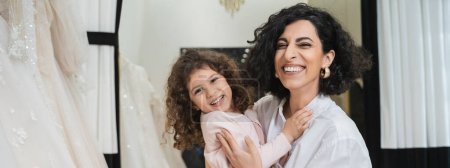 curly little girl hugging cheerful middle eastern bride with brunette hair in white shirt sitting near white wedding dresses in bridal salon, mother and daughter, bonding, special moment, banner 