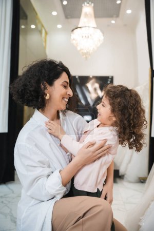 Photo for Cheerful middle eastern bride with brunette hair in white shirt hugging happy little girl near white wedding dresses in bridal salon, mother and daughter, bonding, special moment, shopping - Royalty Free Image