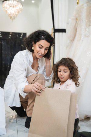 cheerful middle eastern woman with brunette hair in white shirt and surprised little girl looking inside of shopping bag near white wedding dresses in bridal salon, mother and daughter 
