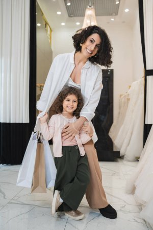 alegre mujer de Oriente Medio con cabello moreno sosteniendo bolsas de compras y abrazando a la linda niña mientras está de pie cerca de los vestidos de novia en el salón de novia, madre e hija, compras nupciales 