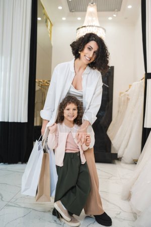 Photo for Joyful middle eastern woman with brunette hair holding shopping bags and hugging cute little girl while standing near white wedding dresses in bridal salon, mother and daughter, bridal shopping - Royalty Free Image