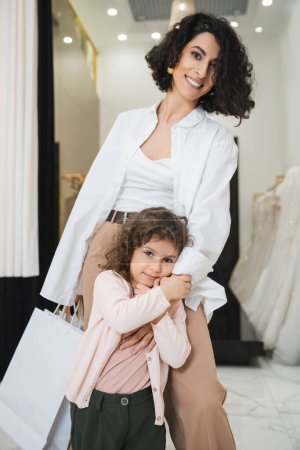 niedliches kleines Mädchen umarmt die Hand einer glücklichen Frau aus dem Nahen Osten mit brünetten Haaren, die Einkaufstüten hält, während sie neben Brautkleidern im Brautsalon steht, Mutter und Tochter, Brauteinkäufe 