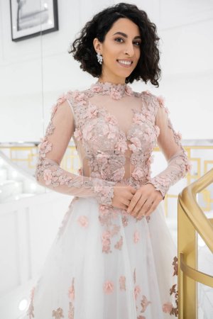 cheerful and beautiful middle eastern bride with brunette and wavy hair standing in gorgeous floral wedding dress and looking at camera inside of bridal boutique, golden accents, luxurious 
