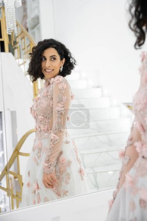 delightful and beautiful middle eastern bride with brunette and wavy hair standing in gorgeous floral wedding dress and looking at mirror inside of bridal boutique, golden accents, luxurious 
