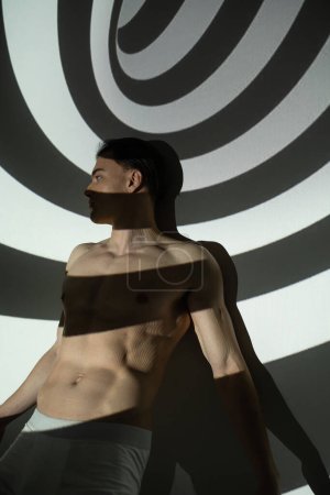 junger, sexy und charismatischer Mann mit hemdlosem Körper, muskulösem Oberkörper, in Unterhosen, die im Stehen wegschauen und auf abstraktem Schwarz-Weiß-Hintergrund mit Spiralprojektion posieren