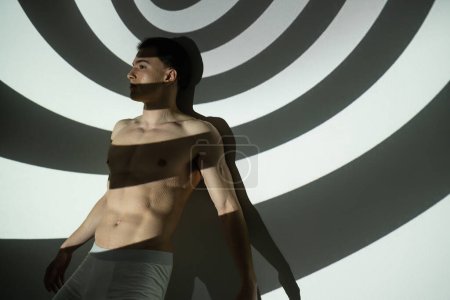 junger, sexy und provokanter Mann mit hemdlosem Oberkörper und muskulösem Körper, der in Unterhosen posiert, während er auf geheimnisvollem schwarz-weißem Hintergrund mit Spiralprojektion steht und wegschaut