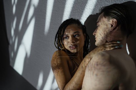 passionate african american woman with dreadlocks embracing young and shirtless man while seducing him on white textured background with white shadows, intimate moment of interracial couple
