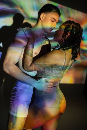 intimate moment of shirtless and muscular man embracing tattooed african american woman with dreadlocks, in net bodysuit on black background with colorful projection and lighting effects