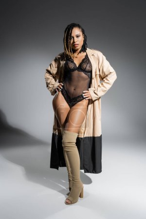 femme afro-américaine pleine longueur avec dreadlocks, en manteau beige à la mode, body sexy en dentelle noire et bottes au-dessus du genou debout avec les mains sur les hanches et en regardant la caméra sur fond gris