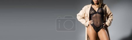 Photo for Partial view of graceful and sexy african american woman in fashionable beige trench coat and black lace lingerie posing with hands on hips on grey background, banner - Royalty Free Image