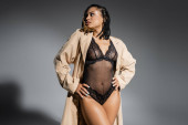 expressive african american woman with dreadlocks, in sexy black lace bodysuit and stylish beige trench coat posing with hands on hips and looking away on grey background Longsleeve T-shirt #658773532