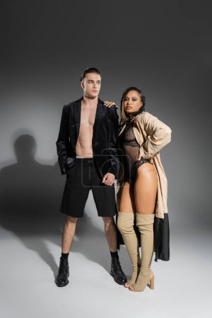 Photo for Full length of interracial couple, trendy man in black silk blazer and shorts, african american woman in lace bodysuit, beige trench coat and over knee boots posing on grey background with shadows - Royalty Free Image