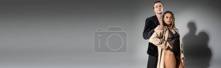 Foto de Sexy mujer afroamericana en traje de encaje y gabardina beige posando con la mano en la cadera cerca de hombre guapo en chaqueta de seda negro sobre fondo gris, bandera - Imagen libre de derechos