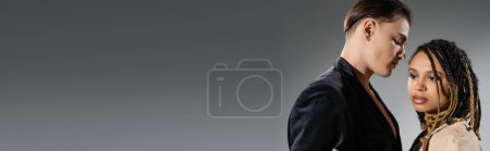 Foto de Joven y cautivante mujer afroamericana con rastas con estilo mirando hacia otro lado cerca de hombre de moda en chaqueta de seda negro sobre fondo gris, bandera - Imagen libre de derechos