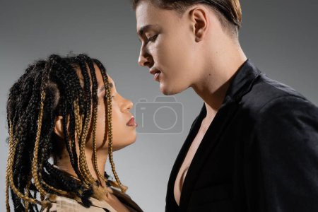 Foto de Vista lateral de pareja interracial sexy, mujer afroamericana con rastas elegantes y joven hombre guapo usando chaqueta de seda negra sobre fondo gris - Imagen libre de derechos