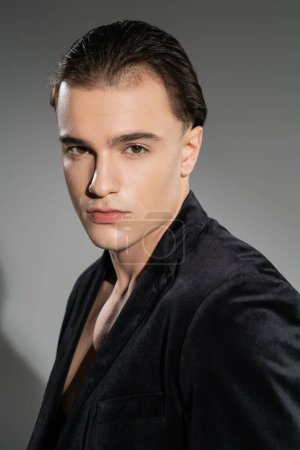 portrait of young and stylish man with confident face expression and brunette hair looking at camera while posing in black silk blazer on grey background