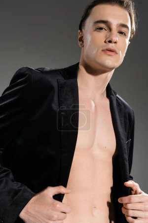 low angle view of provocative and young good looking man wearing black silk blazer on shirtless body while standing and looking at camera on grey background
