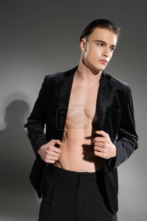 junger, selbstbewusster und modischer Mann mit brünetten Haaren posiert in schwarzem und seidenem Blazer auf schulterfreiem muskulösem Körper und schaut vor grauem Hintergrund weg